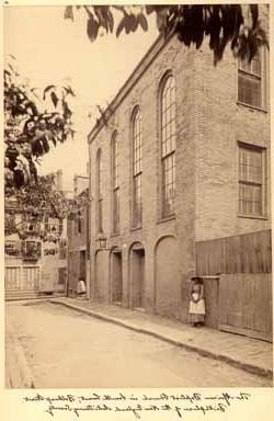 <p>Sepia photograph of a street and the side of a tall, 砖房，有四扇拱门和一盏大铁灯. 在大楼的左边, 披着披肩的黑人妇女, 帽子, 围裙, 她的长袖连衣裙靠墙站着，双手紧握在胸前.</p>